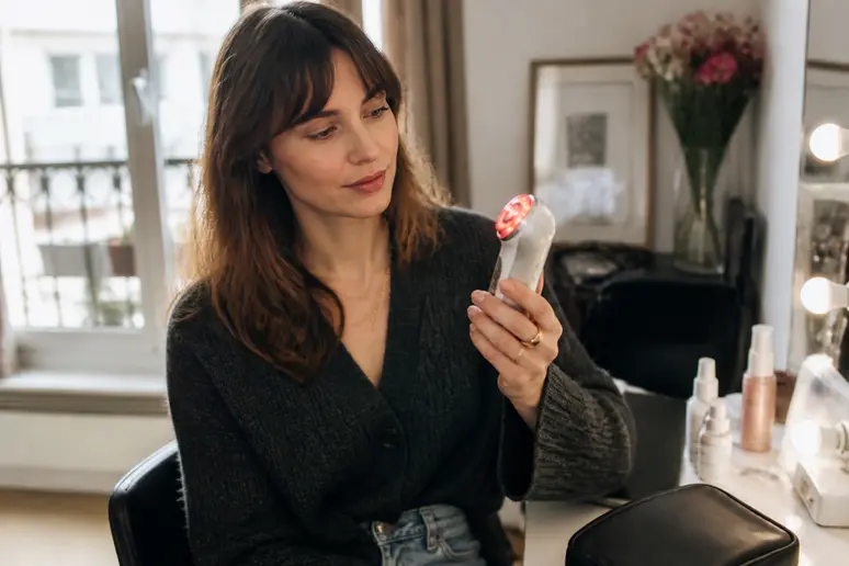 Une femme parisienne examine attentivement un appareil de soin de la peau doté de lumières LED devant son miroir.