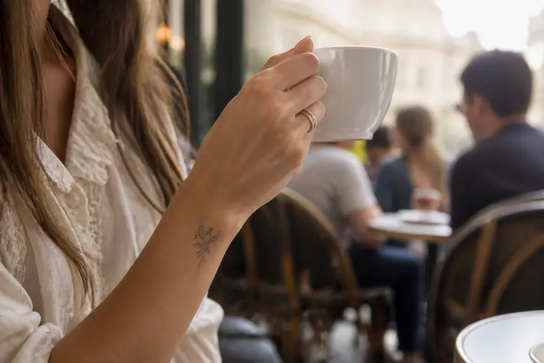 Jeune femme montrant discrètement un tatouage phénix délicat sur son poignet dans un café parisien lumineux, tenant une tasse.