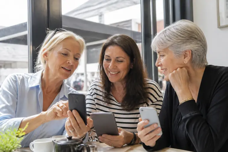 Trois femmes françaises d'un certain âge échangent des avis positifs sur des produits de beauté Luma Cosmetics dans un café parisien ensoleillé.