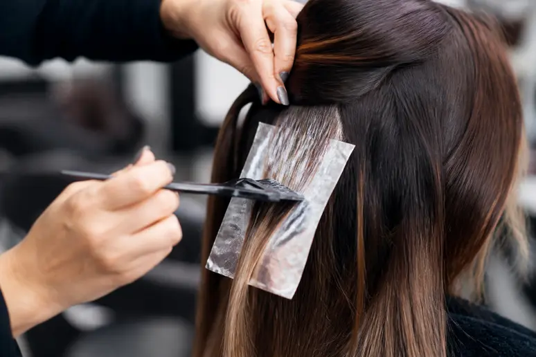 Les mains d'une coiffeuse appliquent avec subtilité des mèches miel et caramel pour un effet balayage naturel et fondu.